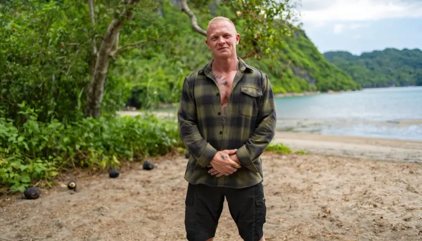 Alexander Strandberg Robinson står på en strand med gröna kullar och vatten i bakgrunden.