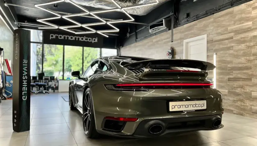 Ciemnozielone Porsche 911 w garażu z nowoczesnym oświetleniem i stojącym obok pionowym banerem.