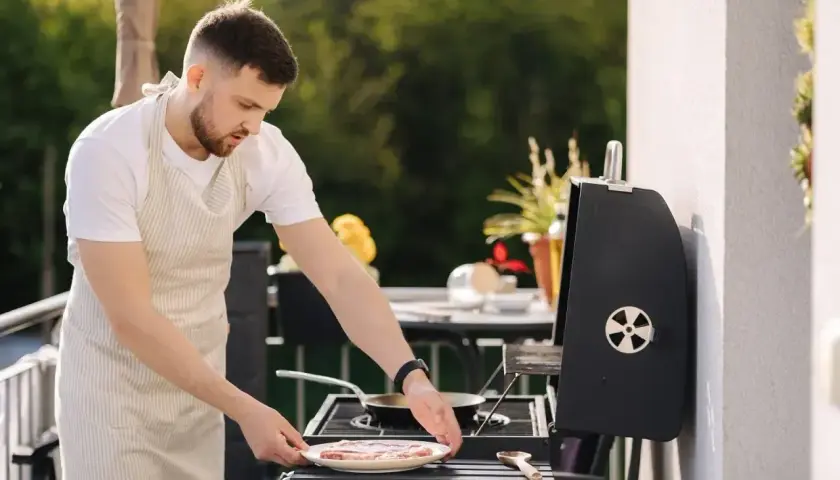Mężczyzna w fartuchu przygotowuje mięso na grillu na balkonie.