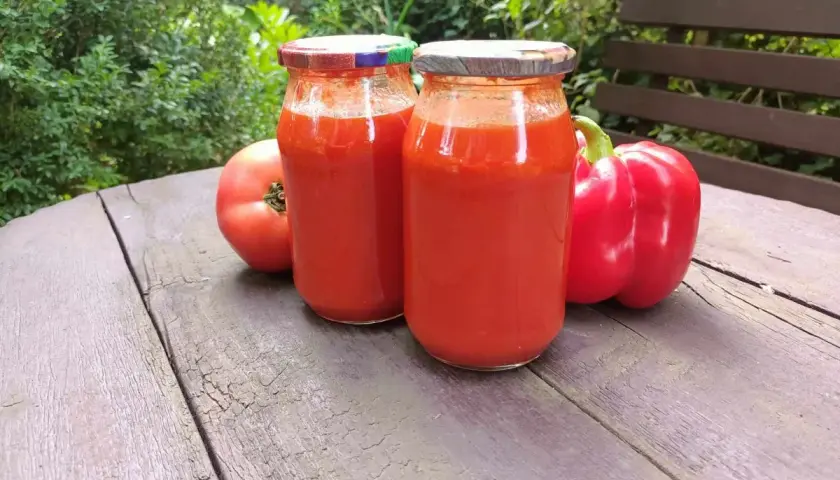 Dwa słoiki domowego sosu paprykowo pomidorowego, obok świeże pomidory i czerwona papryka.