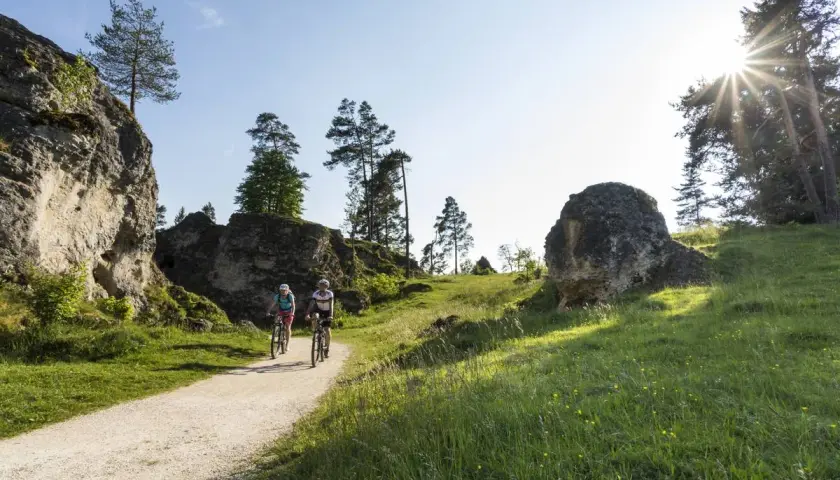 Zwei Radfahrer genießen eine Tour durch eine felsige Landschaft. Sie fahren auf einem Schotterweg, umgeben von grünen Wiesen und Bäumen.