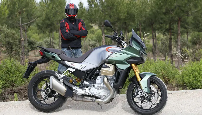 Motociclista con casco posa junto a una moto Guzzi Mandello verde y plateada, lista para la aventura en un camino rural.