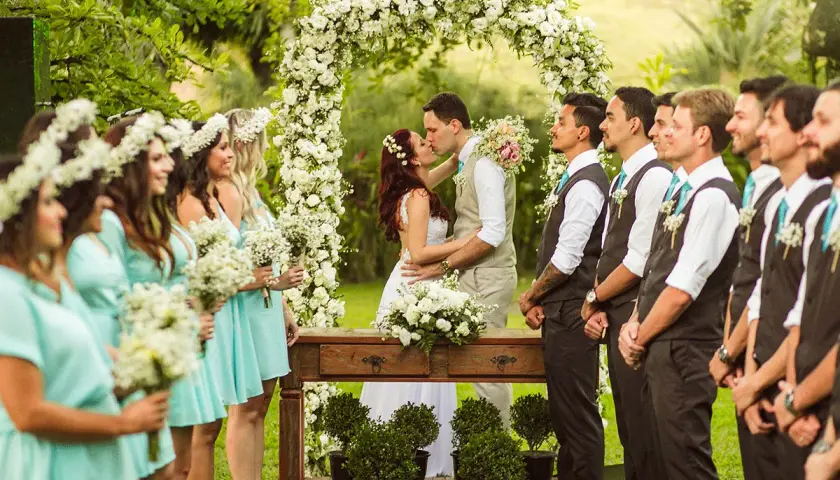 Novios besándose bajo un arco de flores, rodeados de damas de honor y padrinos.