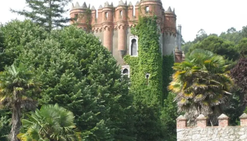 El **palacio de Ocharan**, cubierto de hiedra, se alza entre exuberante vegetación y palmeras, con un muro de piedra en primer plano.