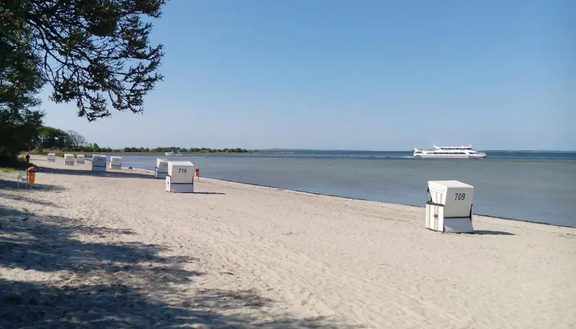 Sonniger Schaprode Strand mit Strandkörben und einem Ausflugsschiff auf dem ruhigen Meer.