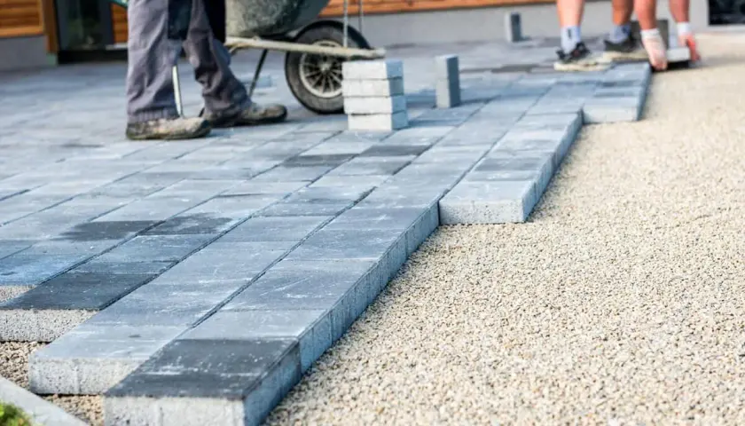 Układanie kostki brukowej na podbudowie z kruszywa. Widać pracowników i taczkę, przygotowanie podłoża to klucz do trwałości, jaki beton pod kostkę jest tu niepotrzebny.