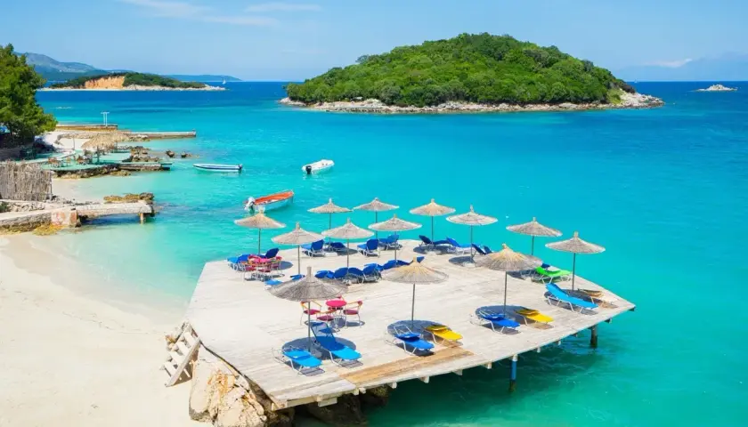 Idealne miejsce, gdzie najlepiej nad morze: piaszczysta plaża, turkusowa woda, drewniany pomost z leżakami i parasolami, zielona wyspa w oddali.