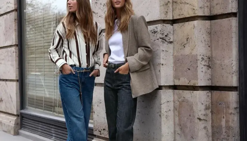 Deux femmes posent devant un mur en pierre. L'une porte un jean tapered femme, une chemise rayée et une ceinture chaîne. L'autre, un blazer et un jean.