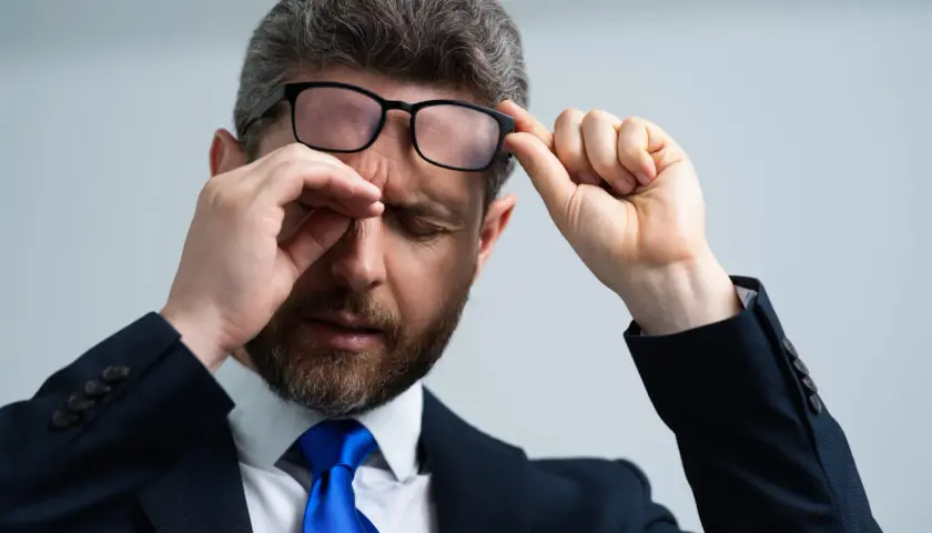 Mężczyzna w garniturze i okularach pociera oko, jakby coś wpadło do oka pod górną powiekę.