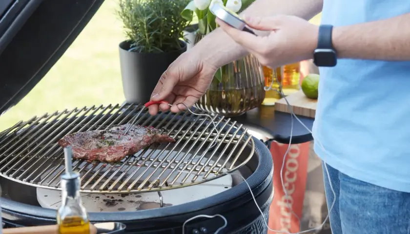 Un homme utilise un thermomètre pour vérifier la cuisson d'un steak sur un barbecue. Le steak est assaisonné et repose sur la grille.