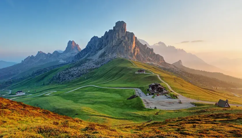 A winding road leads to a mountain lodge, perfect for hiking the Dolomites, Italy. Lush green hills meet jagged peaks under a soft sunrise.