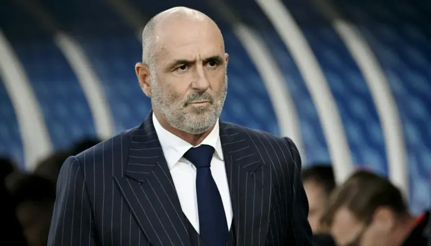 Łysy mężczyzna z brodą, ubrany w granatowy garnitur w paski i białą koszulę, to probierz trener.