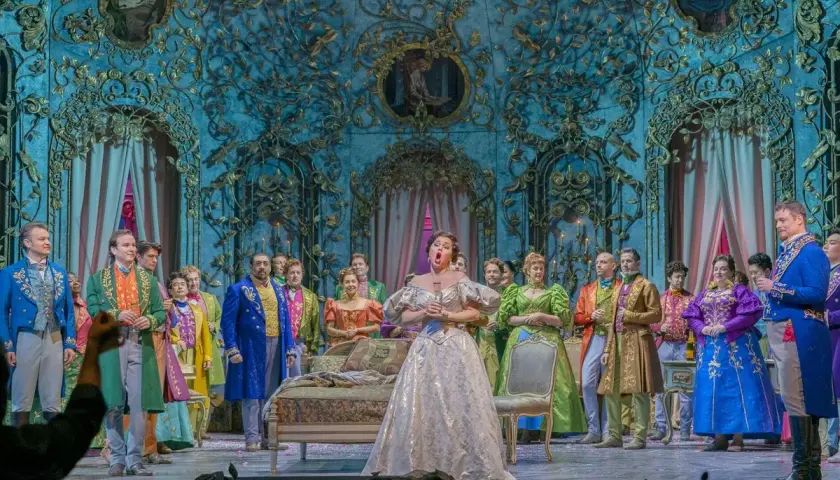 Primadonna w lśniącej sukni śpiewa na scenie operowej, otoczona przez barwnie ubranych artystów.