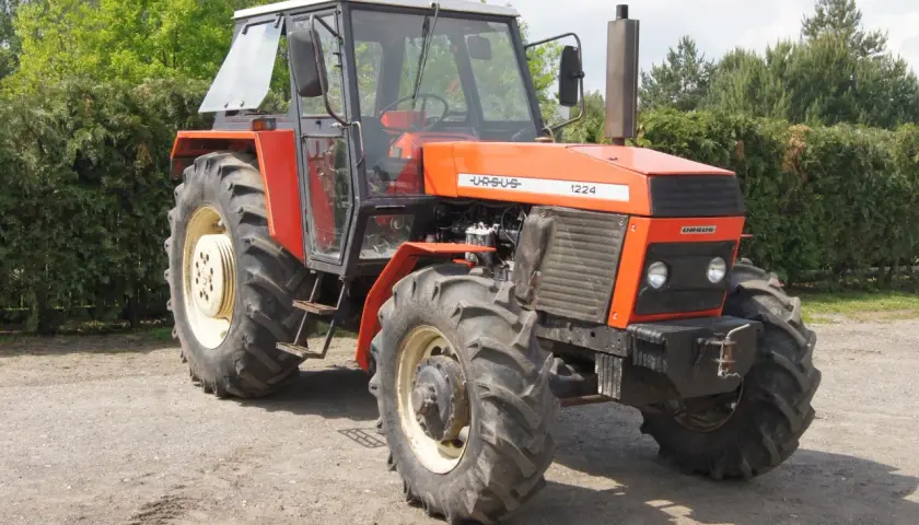 Pomarańczowy Ursus 1224 z kabiną stoi na drodze gruntowej, otoczony zielenią drzew i krzewów.