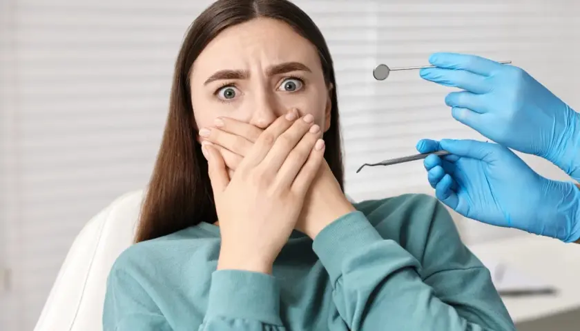 Przerażona pacjentka z dentofobią zakrywa usta. W pobliżu widać ręce dentysty w niebieskich rękawiczkach z narzędziami.