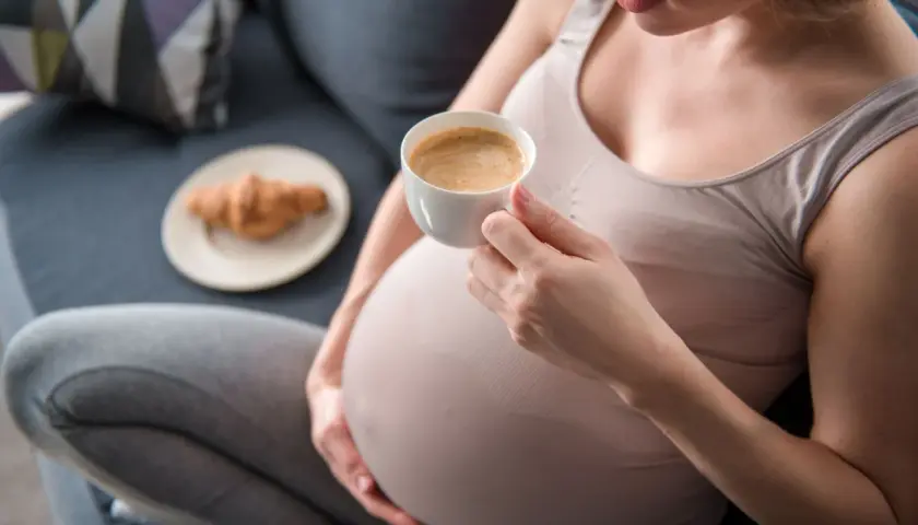 Kobieta w ciąży delektuje się kawą rozpuszczalną, siedząc na kanapie z rogalikiem na talerzyku obok.
