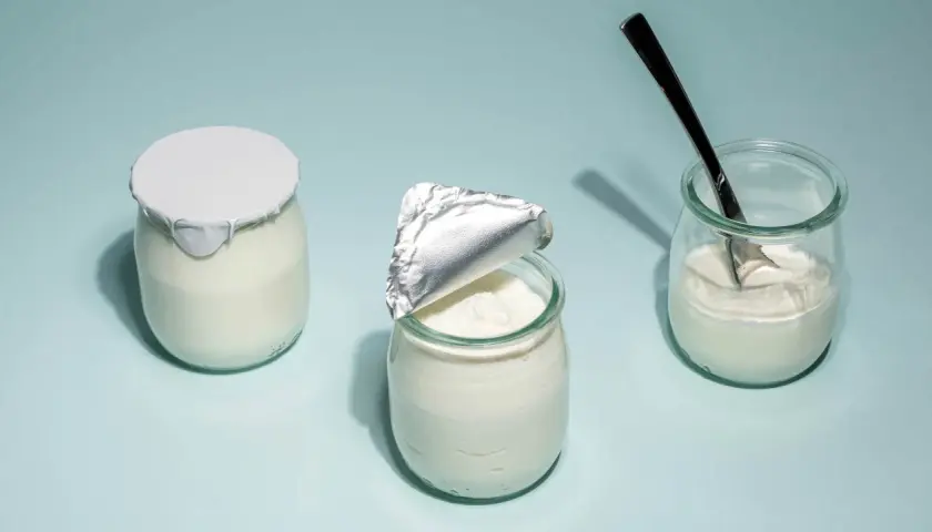Tre burkar med hemmagjord yoghurt på ljusblå bakgrund. En är helt försluten, en annan är delvis öppnad och en tredje har en sked i sig.
