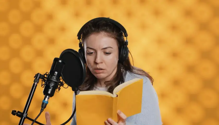 Eine Synchronsprecherin mit Kopfhörern liest ein Buch vor Mikrofon. Sie fragt sich, wie hoch wohl ihr Synchronsprecher Gehalt sein wird.