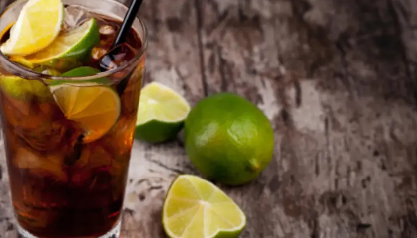Cuba Libre mit dunklem Rum, Eis und Limettenspalten auf Holzhintergrund.