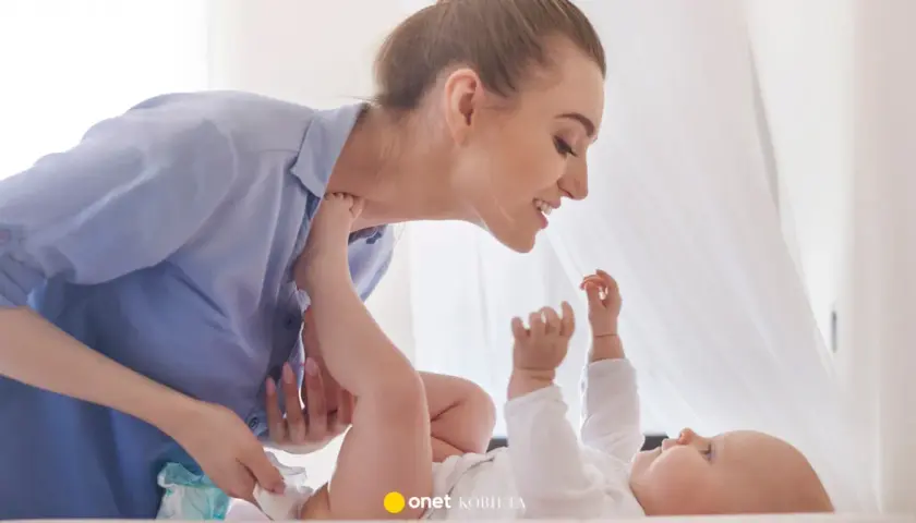 Uśmiechnięta mama pochyla się nad dzieckiem, które leży na plecach. Wykonuje szerokie pieluchowanie, delikatnie unosząc nóżki malucha.