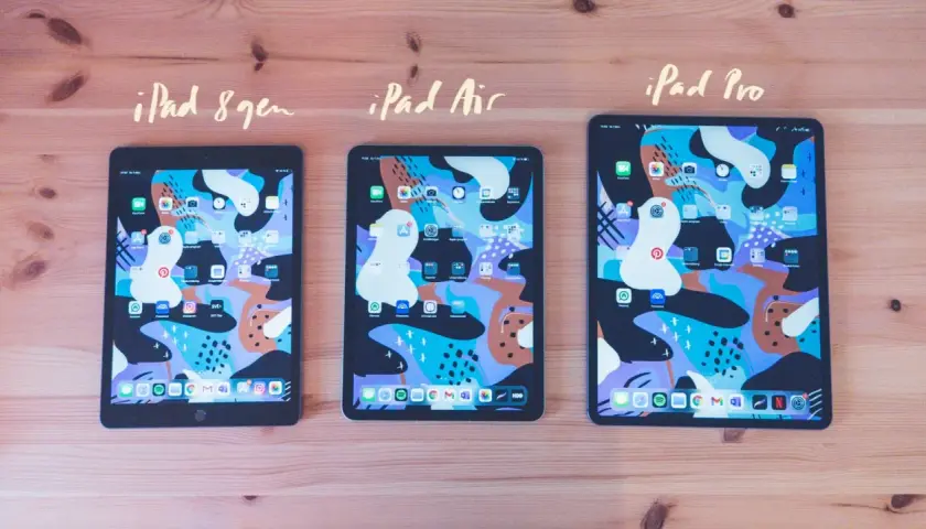 Tre iPads, 8:e gen, Air och Pro, ligger på ett träbord. Alla har samma färgglada bakgrundsbild och appar. Det är lätt att skriva på iPad.