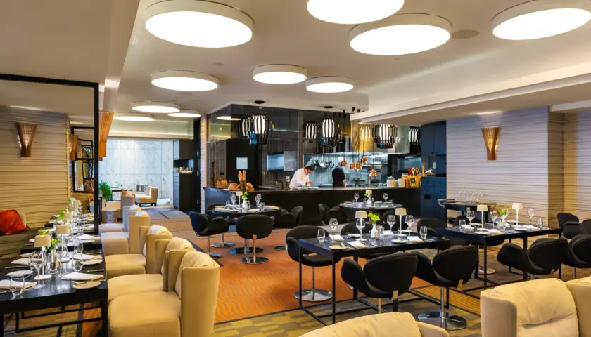 Elegancka brasserie z widokiem na kuchnię, gdzie szef kuchni przygotowuje dania. Stoliki nakryte do kolacji, wygodne fotele i nowoczesne oświetlenie tworzą przytulną atmosferę.