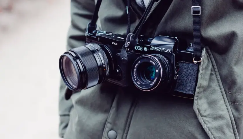 Osoba w zielonej kurtce nosi na szyi dwa aparaty fotograficzne: jeden cyfrowy, drugi analogowy.