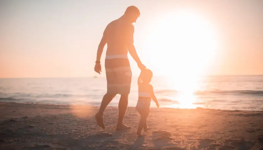 Ojciec prowadzi za rękę swoje dziecko 4 lata, spacerując po plaży o zachodzie słońca.