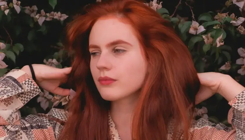 Chica con vibrante pelo pelirrojo, rodeada de flores y follaje, con las manos jugando con su cabello.