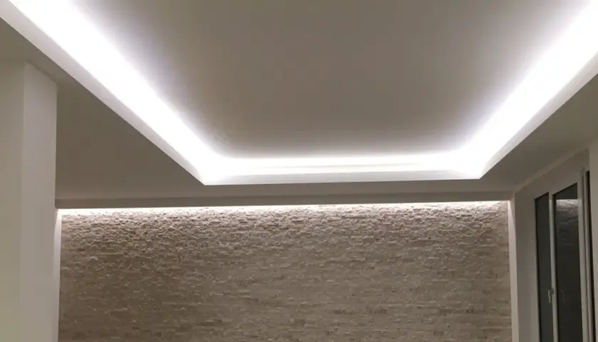 Illuminazione indiretta su velette in cartongesso, che creano un effetto soffuso su una parete in pietra chiara.
