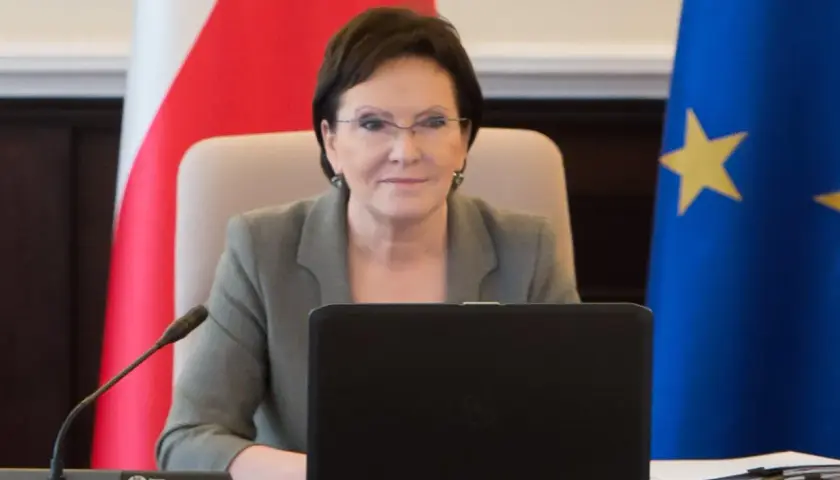 Ewa Kopacz, premier, siedzi przy stole z laptopem, obok flagi Polski i Unii Europejskiej.
