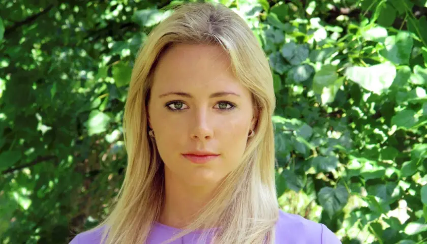 Close-up portrait of a blonde woman with green eyes and a purple top, against a blurred green foliage background.