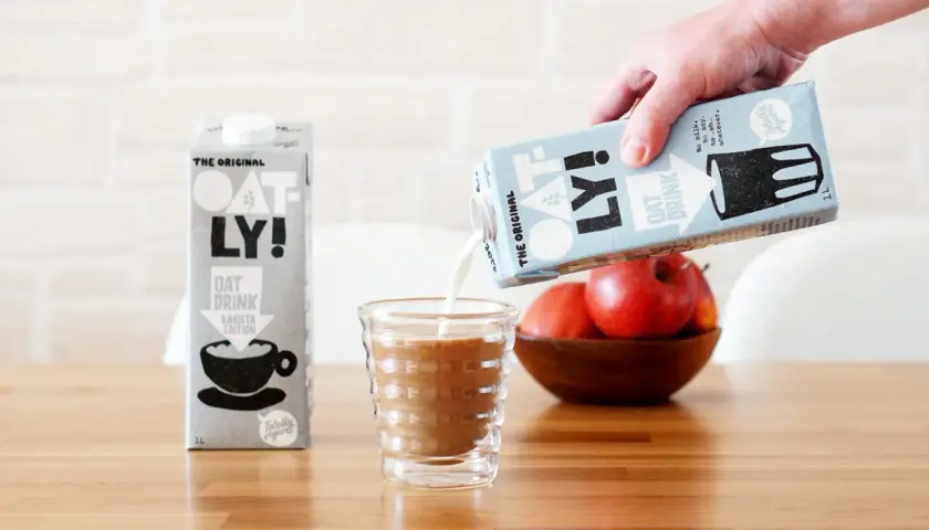 Ręka wylewa mleko dla baristów Oatly do szklanki. Obok stoi karton tego samego napoju.