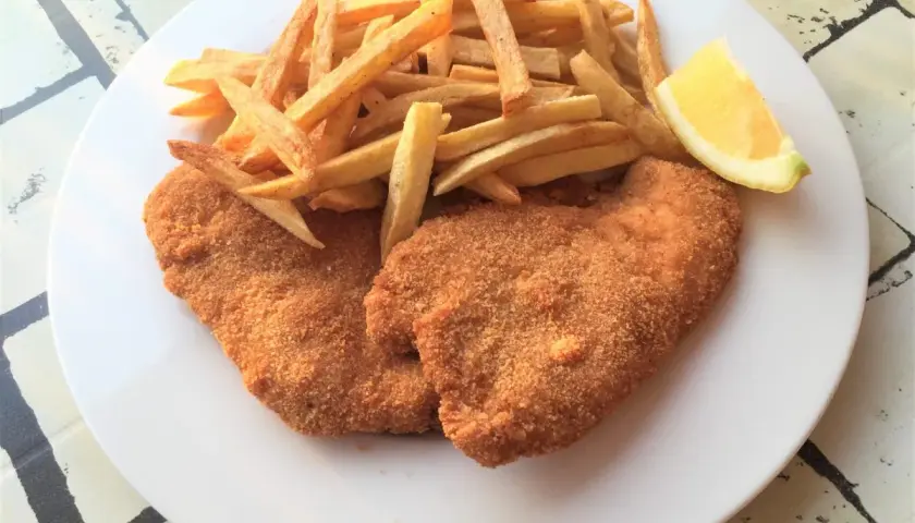 Due cotolette di pollo dorate e croccanti, accompagnate da patatine fritte e uno spicchio di limone su un piatto bianco.