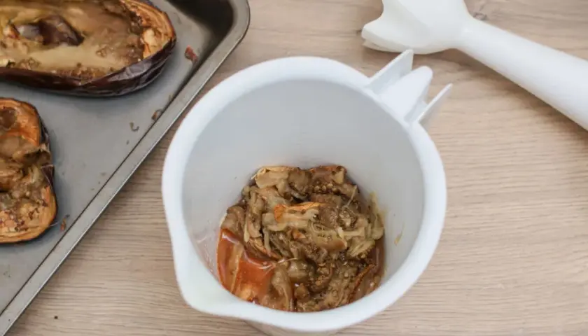 Berenjenas asadas en una bandeja, listas para convertirse en un delicioso hummus de berenjena. Un batidor de mano espera para mezclar.