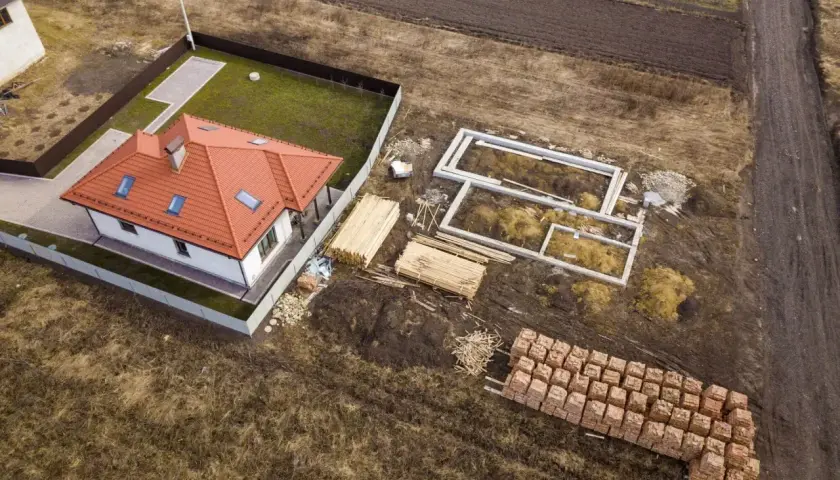 Zabudowa zagrodowa w budowie: gotowy dom z czerwonym dachem obok fundamentów i materiałów budowlanych.
