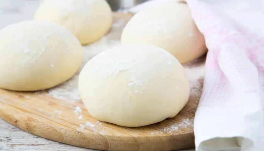 Kulki ciasta, idealne ciasto na pizzę, posypane mąką, leżą na desce. Obok różowy ręcznik.