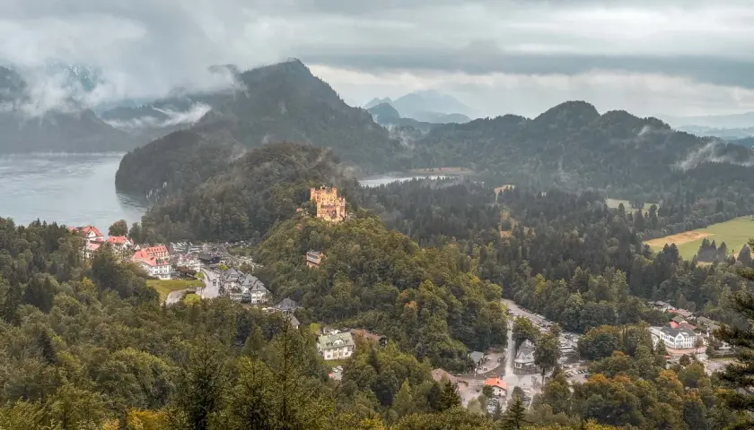 Schlechtes Wetter trübt den Urlaub. Ein gelbes Schloss thront auf einem bewaldeten Hügel über einem Dorf am See.