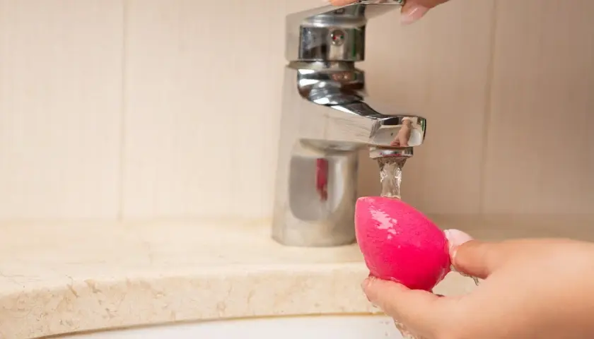 Mano limpiando esponja de maquillaje rosa bajo un grifo. Aprende como limpiar las esponjas de maquillaje para un mejor acabado.