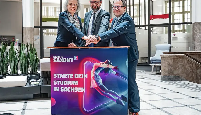 Drei Personen legen ihre Hände auf ein Banner für ein berufsbegleitendes Studium in Leipzig.