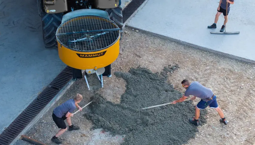 Zwei Arbeiter verteilen frischen Beton, der aus einem gelben Zwangsmischer einer Mammut-Maschine fließt. Ein dritter Arbeiter glättet den Beton.