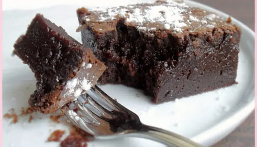 Un morceau de gâteau chocolat, saupoudré de sucre glace, est sur une assiette blanche. Une fourchette est prête à déguster ce délice.