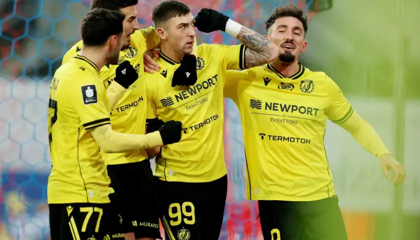 Piłkarze w żółtych koszulkach świętują bramkę. Fani spekulują o plotkach transferowych Widzewa, a drużyna cieszy się sukcesem.