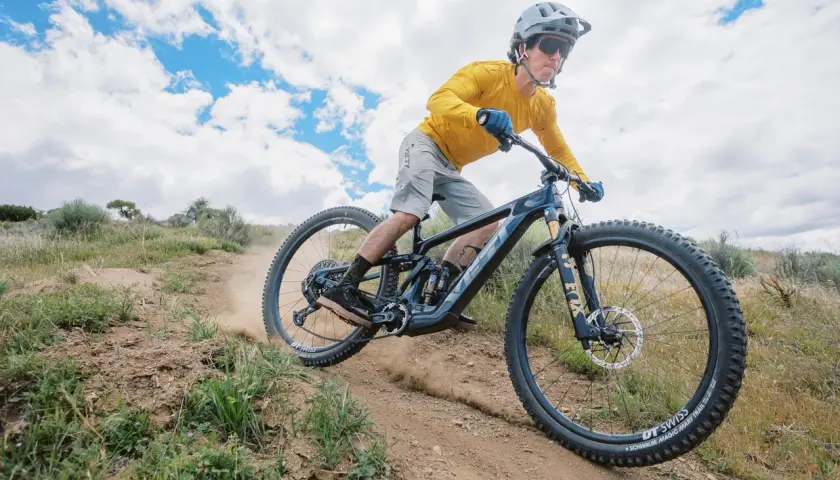 A mountain biker shreds a dusty trail on a Yeti MTE bike, kicking up dirt under a cloudy sky.