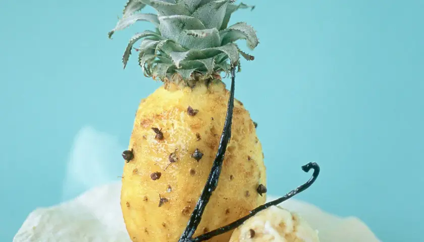 Un ananas rôti, parsemé de clous de girofle et de gousses de vanille, sur un fond bleu pâle. Un délice sucré et épicé.