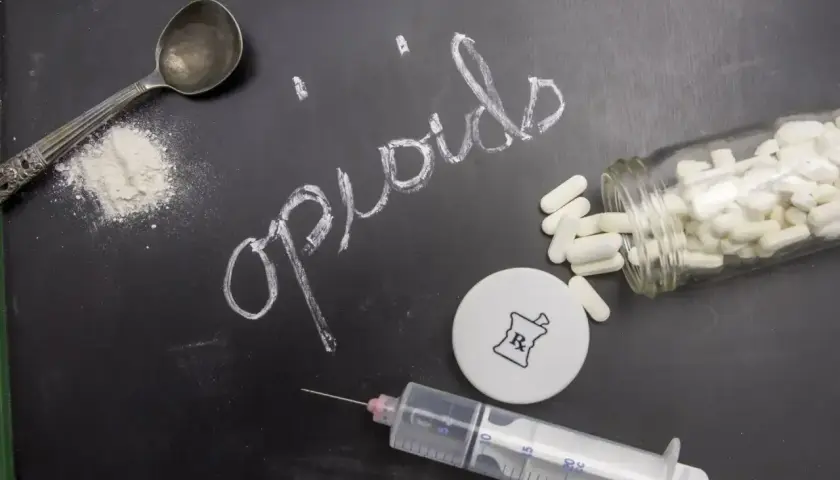 Na czarnej tablicy napis "opioids" kredą. Obok łyżka z proszkiem, strzykawka, fiolka z kapsułkami i symbol apteczny.