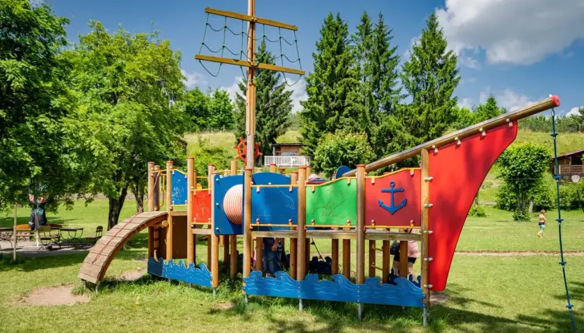 Kolorowy plac zabaw w kształcie statku pirackiego, z drabinkami i zjeżdżalnią, to wspaniałe atrakcje dla dzieci nad brzegiem jeziora.