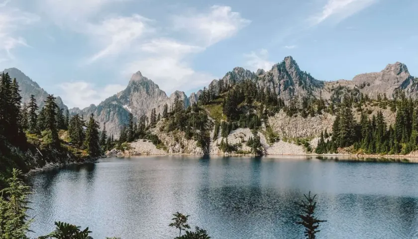 A serene alpine lake reflects jagged, tree-covered mountains. This view makes you wonder how long to hike 16 miles to reach such a peaceful spot.