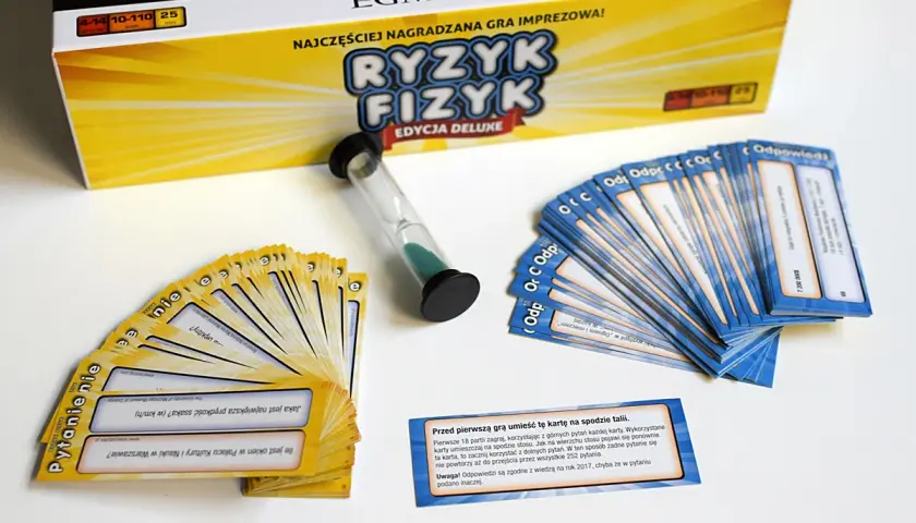 Pudełko gry "Ryzyk Fizyk" i rozłożone karty pytań i odpowiedzi. Czasomierz piaskowy czeka na rozpoczęcie rundy.