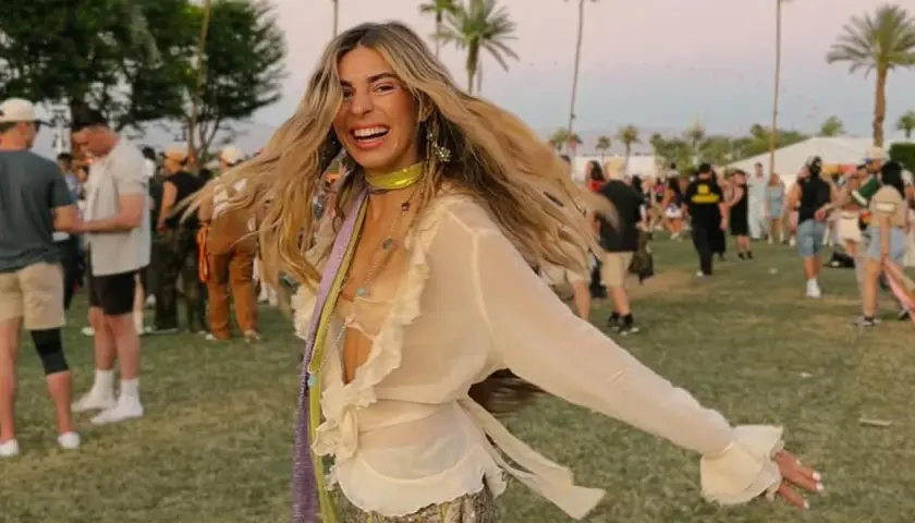 Mujer sonriente y con el pelo al viento disfruta de un look festival verano, rodeada de palmeras y gente.